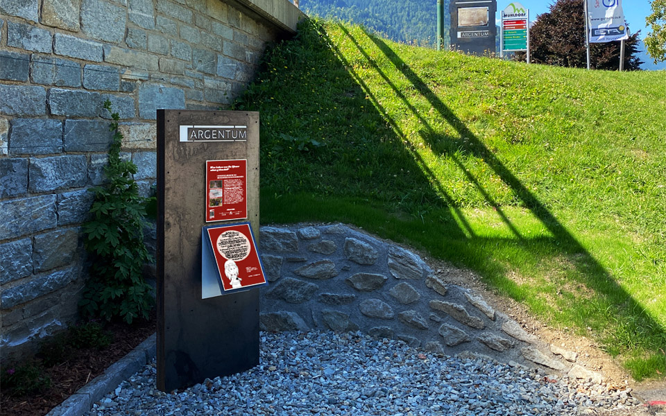 The picture shows a station on the Roman path in Mühldorf. In the foreground is a metal surface on which two plaques are installed. On the metal surface, at eye level for adults, there is a plaque with an informative text about the history of Mühldorf. Directly below it, also at children's eye level, is a second panel. This offers a child-friendly text on the topic. Behind the metal wall is a stone wall, on the right-hand side of the picture you can see an embankment.