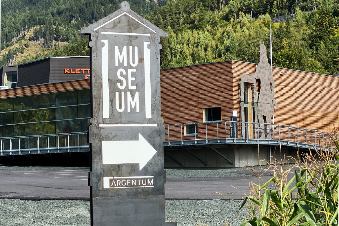 In the foreground of the picture is a tower-like sculpture that serves as a guiding element. Museum Argentum is engraved on this sculpture and an arrow points to the right in the direction of the museum entrance. In the middle ground of the picture, the Museum Argentum presents itself with a wooden façade. An imposing sculpture depicting a Celtic warrior stands right next to the entrance to the museum. The warrior towers above the building. A wooded mountain stretches out in the background.