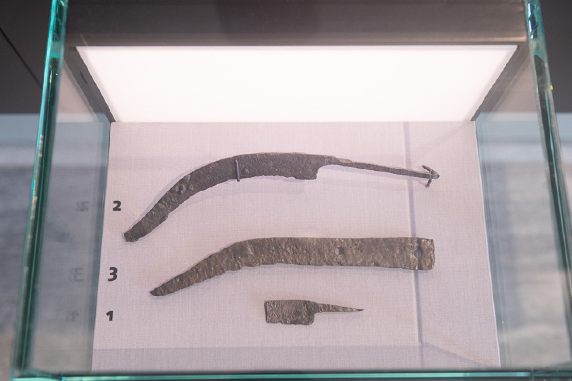 Das Bild zeigt ein Ausstellungsstück im Museum ARGENTUM: eine vermutlich antike Sichel. Das Exponat ist hinter einer klaren Glasscheibe gesichert, um es vor Berührung und Umwelteinflüssen zu schützen.