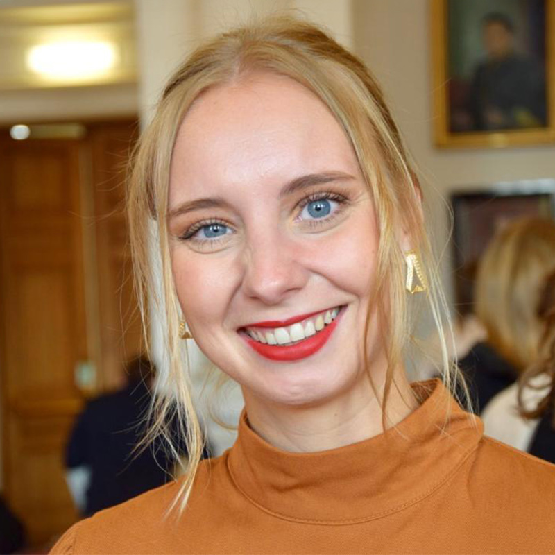 The picture shows Laura Lucia Pösendorfer, MA BA, in her role as deputy curator at the ARGENTUM Museum. Her blonde hair is elegantly tied back, with a few strands falling out of the braid and gently cascading downwards. Laura wears earrings and an elegant sweater in a warm color palette between orange and brown