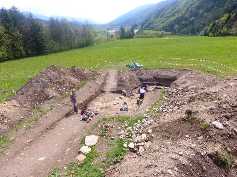 The picture, taken from a bird's eye view, shows an archaeological excavation in the Mühldorf area in the foreground. Three people can be seen in the picture who are involved in excavation work.