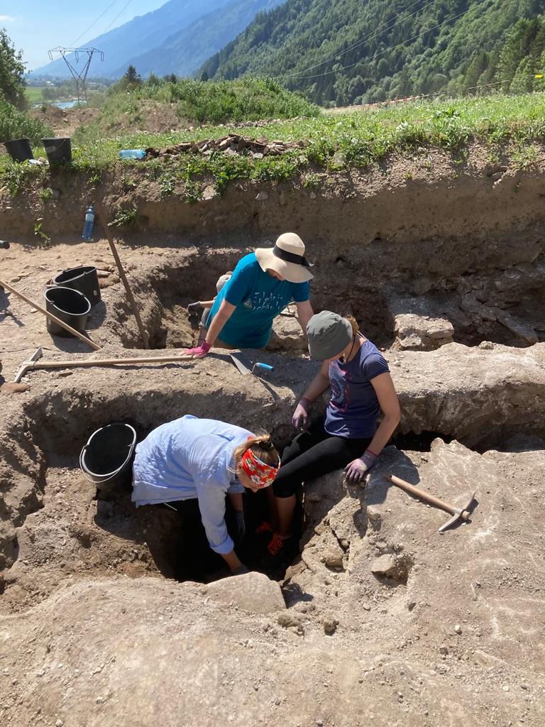 Auf dem Bild befinden sich drei Personen, die sich in hüfthohen, ausgegrabenen Gängen befinden, während sie nach alten Schätzen suchen. Um sie herum liegen Eimer und andere Grabungswerkzeuge.