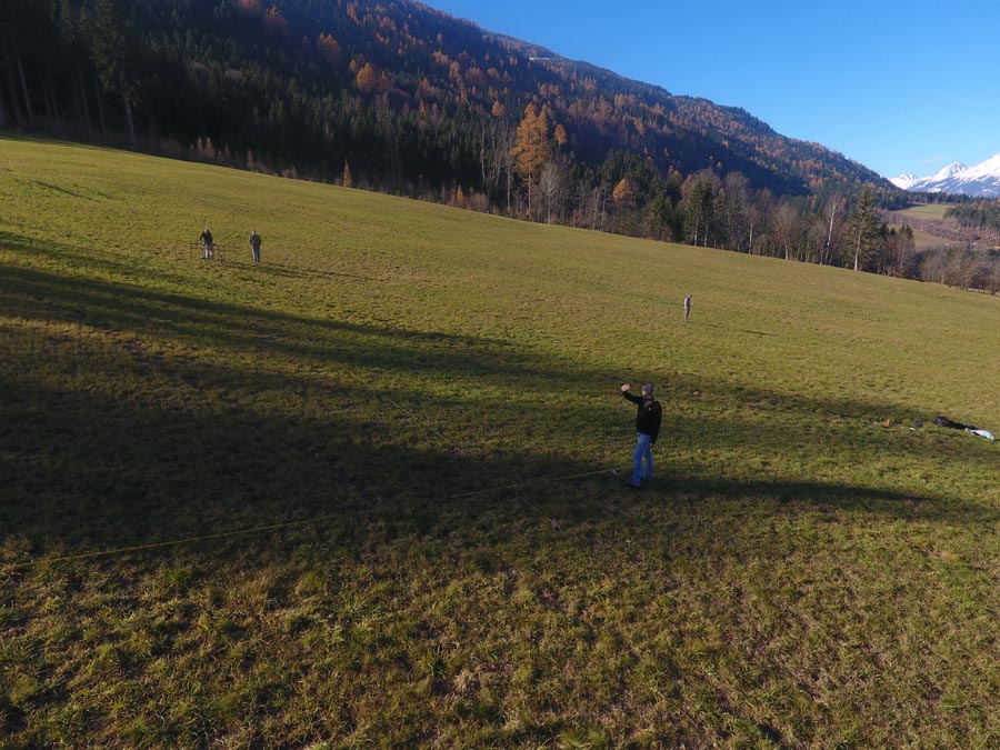 The picture shows a large meadow on which four people are spread out. The person is carrying out ground penetrating radar measurements. In the background is a dense forest. There is a mountain above the forest.
