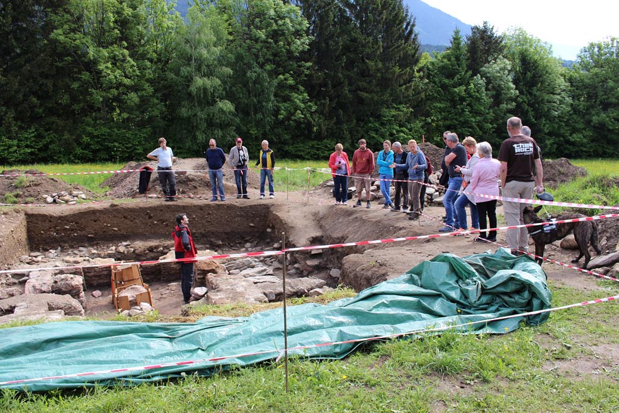 The picture shows a guided tour of an archaeological excavation site. The lecturer is deep in the excavation. About 15 listeners stand in a group and listen to the words of the lecturer. The excavation site itself is secured by barriers. The audience stands behind it. A forest can be seen in the background.