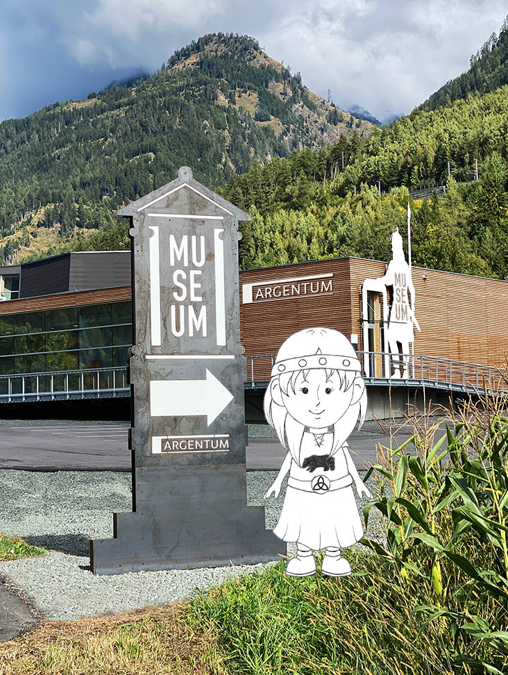 In the foreground of the picture is a tower-like sculpture that serves as a guiding element. Museum Argentum is engraved on this sculpture and an arrow points to the right in the direction of the museum entrance. Right next to the sculpture is the Celtic girl Matugenta, who has been photoshopped into the picture. In the middle ground of the picture is the Argentum Museum with a wooden façade. An imposing sculpture depicting a Celtic warrior stands right next to the entrance to the museum. The warrior towers above the building. A wooded mountain stretches out in the background.